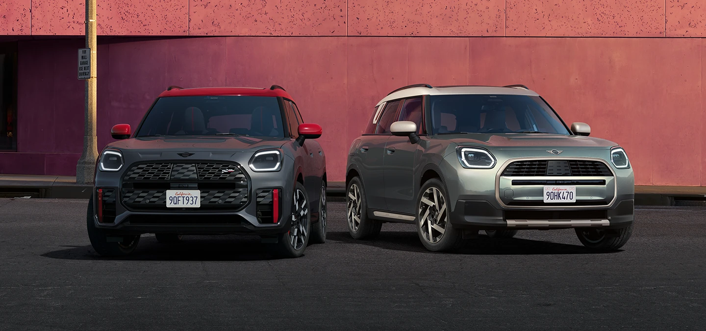 Widok z przodu na MINI Countryman JCW w kolorze Legend Grey z czerwonym dachem oraz MINI Countryman w kolorze Smokey Green z dachem w kolorze Vibrant Silver stojące obok siebie.