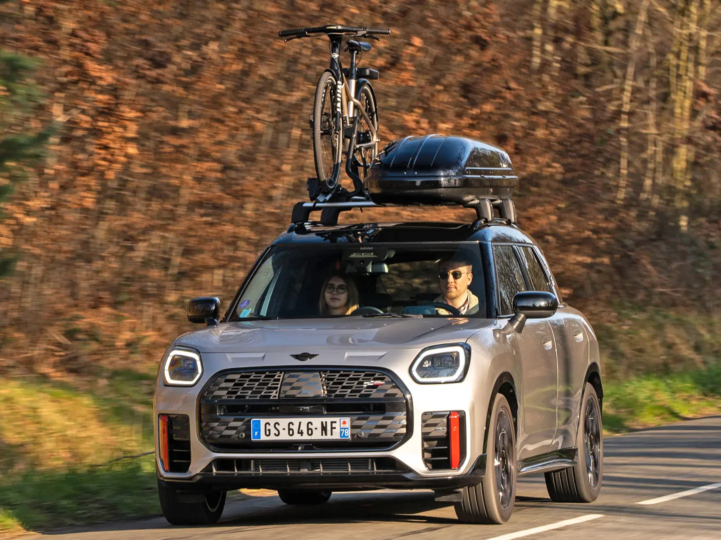 MINI Countryman Cooper w kolorze srebrnym na górskiej drodze z bagażnikiem dachowym MINI, pojemnikiem dachowym MINI, dachowym bagażnikiem rowerowym MINI z jednym zamontowanym rowerem oraz wingletem MINI John Cooper Works – widok z przodu. 