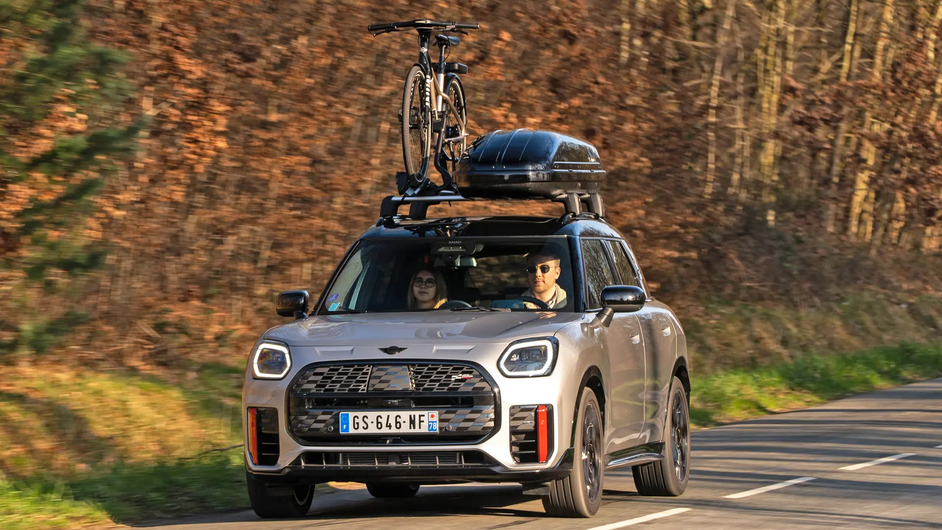 MINI Countryman Cooper w kolorze srebrnym na górskiej drodze z bagażnikiem dachowym MINI, pojemnikiem dachowym MINI, dachowym bagażnikiem rowerowym MINI z jednym zamontowanym rowerem oraz wingletem MINI John Cooper Works – widok z przodu. 