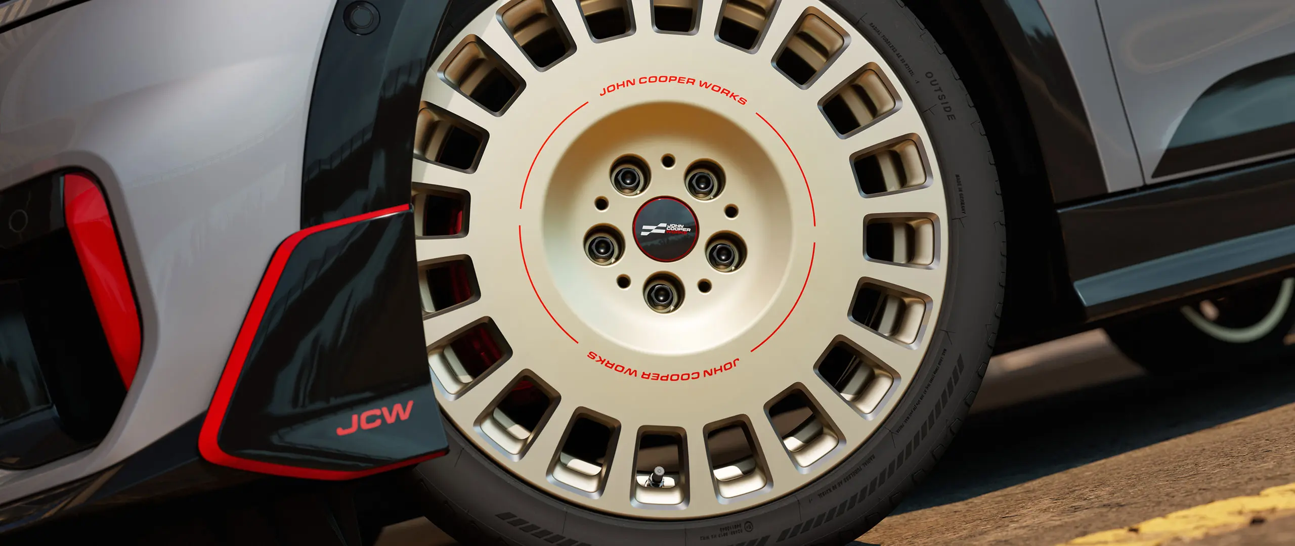 Zbliżenie detali 18-calowej obręczy koła JCW Rallye Spoke 964 w dwukolorowym wykończeniu Vibrant Silver z frezowaniem diamentowym oraz wingletem przednim.