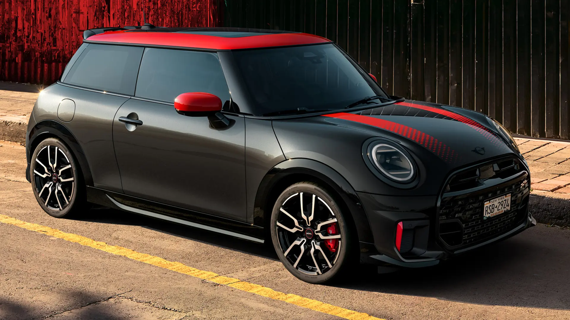 3-drzwiowe MINI John Cooper Works w kolorze Midnight Black z dachem i osłonami lusterek w kolorze Chili Red i czerwonymi paskami sportowymi, widziane z boku i zaparkowane na ulicy.