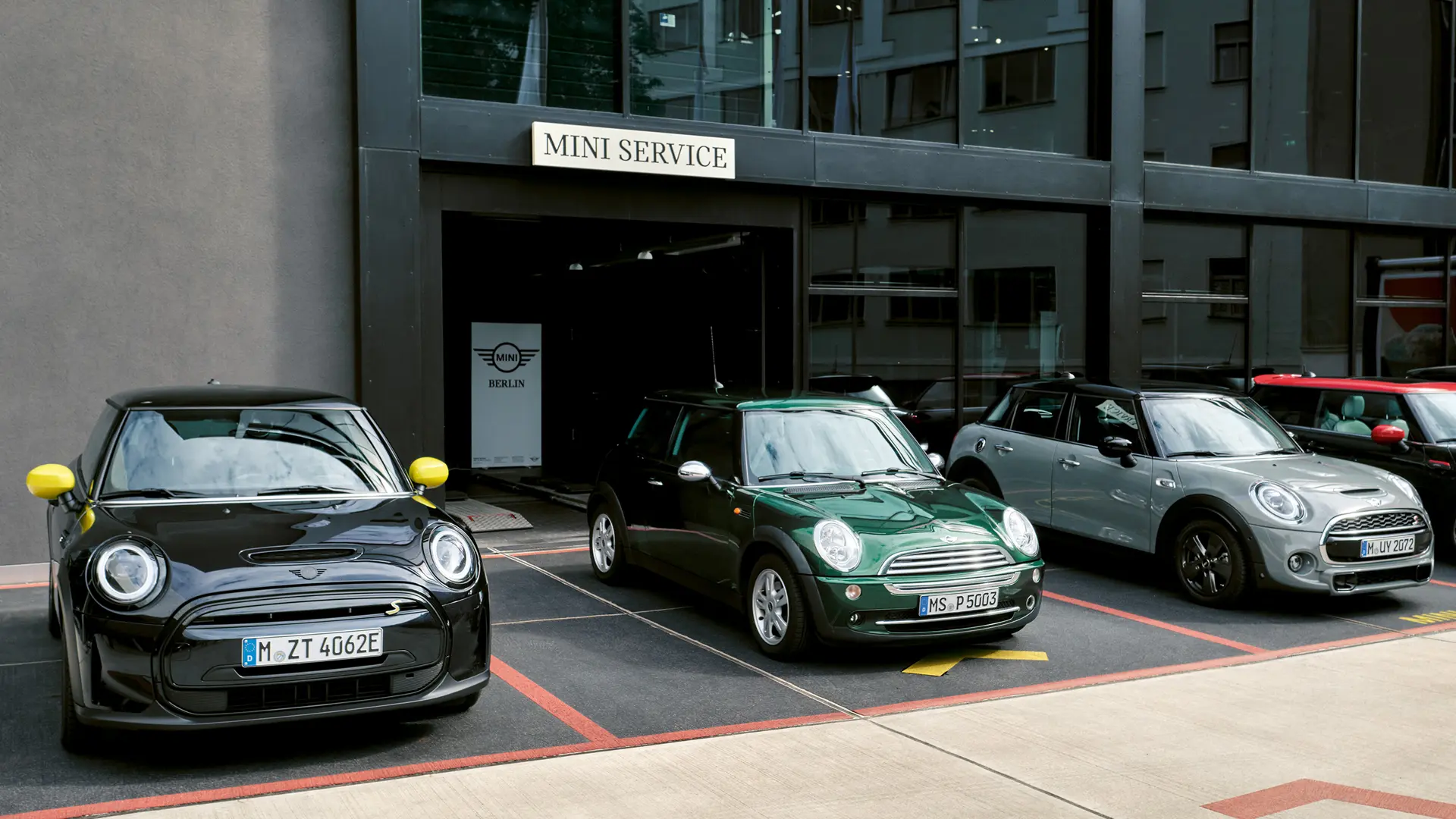 Elektryczne MINI Cooper S w kolorze czarnym stojące przed salonem MINI obok 3-drzwiowego MINI Cooper w kolorze zielonym, 5-drzwiowego MINI w kolorze srebrnym, MINI JCW w kolorze czarnym, widziane z przodu. 