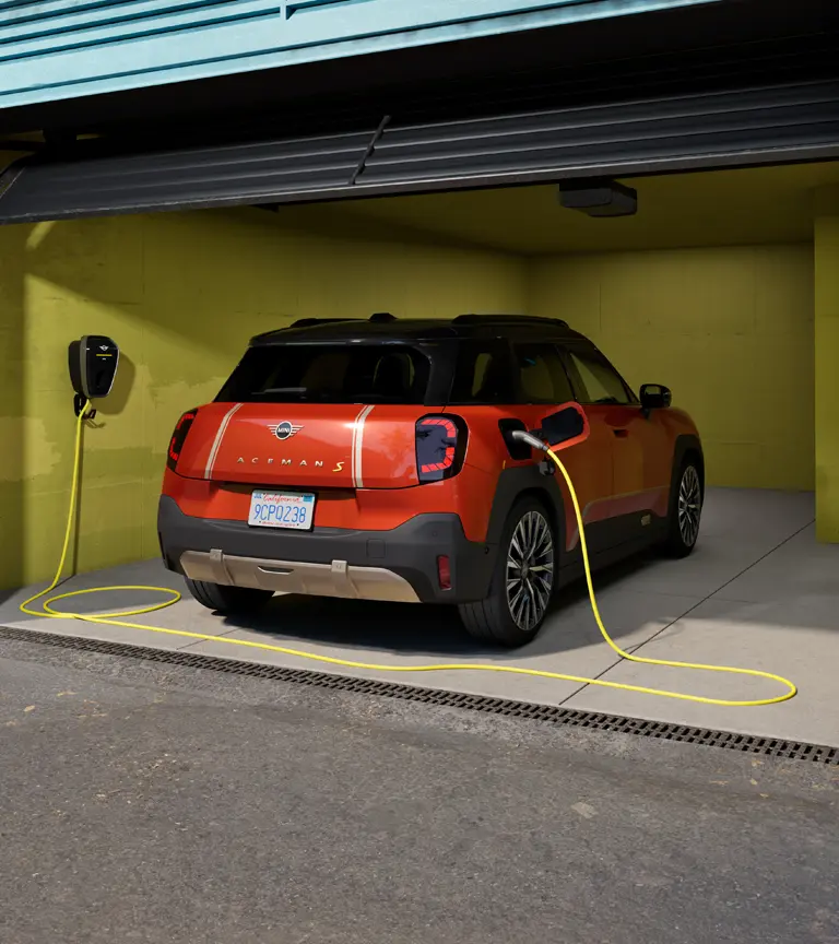 W pełni elektryczne MINI Aceman S ładowane za pomocą stacji ładowania MINI Wallbox Plus, widziane z tyłu, zaparkowane w garażu.