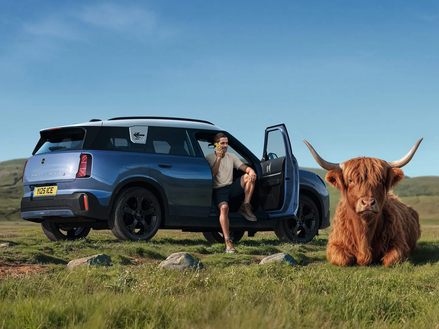 Mężczyzna stoi obok swojego MINI Countryman S i rozmawia z MINI Roadside Assistance za pośrednictwem aplikacji MINI, podczas gdy w pobliżu leży szkocki byk rasy Highland.